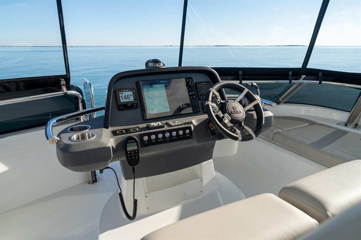 Boat cockpit showing a steering wheel, navigation screen, controls, and open water view with clear skies.